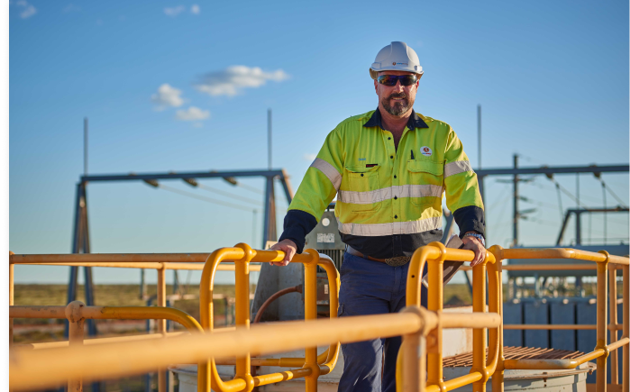 An image of a site worker wearing hi-vis with a hard hat and sunglasses. He is stranding on an elevated platform with a blue sky in the background.